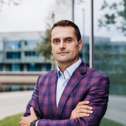 Fotographiki Adam Głodkowski - Portret mężczyzny w bordowej marynarce w kratę, skrzyżowane ręce, smartwatch na nadgarstku, nowoczesny budynek biurowy w tle.