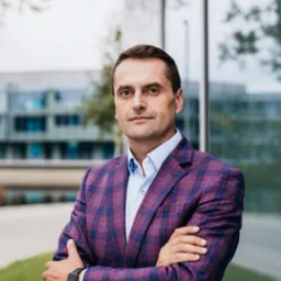 Portret mężczyzny w bordowej marynarce w kratę, skrzyżowane ręce, smartwatch na nadgarstku, nowoczesny budynek biurowy w tle.