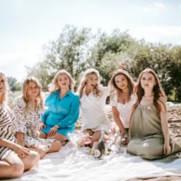 Sześć uśmiechniętych kobiet siedzi na kocu na plaży, obok butelka szampana i kwiaty, w tle drzewa i jezioro.