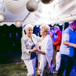 Fotographiki Adam Głodkowski - Dwie uśmiechnięte starsze kobiety tańczące pod białym namiotem z szarymi balonami, widoczni inni goście w tle.