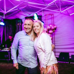 Fotographiki Adam Głodkowski - Para uśmiechnięta i obejmująca się na tle sceny z DJ-em, oświetleniem i dekoracjami w odcieniach fioletu.