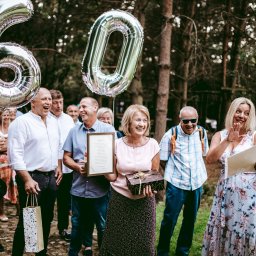 Fotographiki Adam Głodkowski - Uroczystość w plenerze z balonami w kształcie cyfr 60, kobieta trzyma prezent i ramkę z tekstem, a inna osoba gestykuluje, otoczeni przez uśmiechniętych gości na tle drzew.