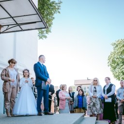 Fotographiki Adam Głodkowski - Grupa osób, w tym dziewczynka w białej sukience i wianku, stoi na schodach przed budynkiem z drewnianymi drzwiami podczas uroczystości.