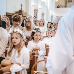 Fotographiki Adam Głodkowski - Uczestnicy Pierwszej Komunii Świętej w kościele, dziewczynki w wiankach trzymają chleb, obecny ksiądz w białej szacie z czerwonym pasem.
