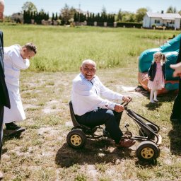 Fotographiki Adam Głodkowski - Uśmiechnięty mężczyzna w białej koszuli jedzie na gokarcie po trawiastym terenie podczas uroczystości, w tle dmuchana zjeżdżalnia, dziecko w różowej sukience i osoby w eleganckich strojach.