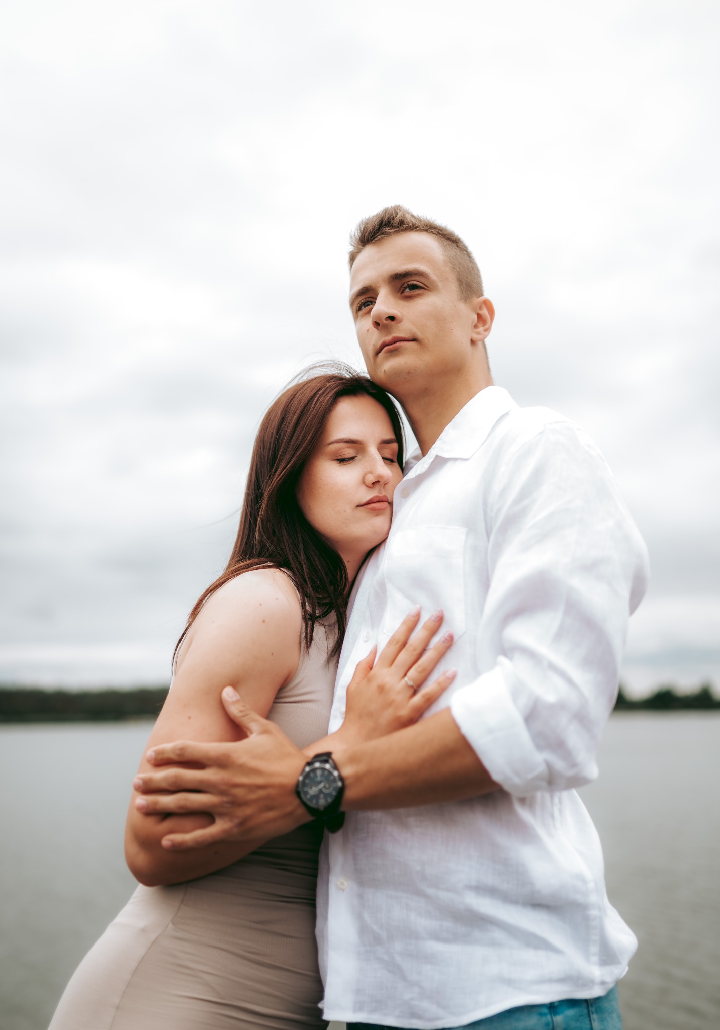 Para zakochanych obejmuje się na tle jeziora i pochmurnego nieba. Mężczyzna w białej koszuli, kobieta w beżowej sukience, z zamkniętymi oczami.