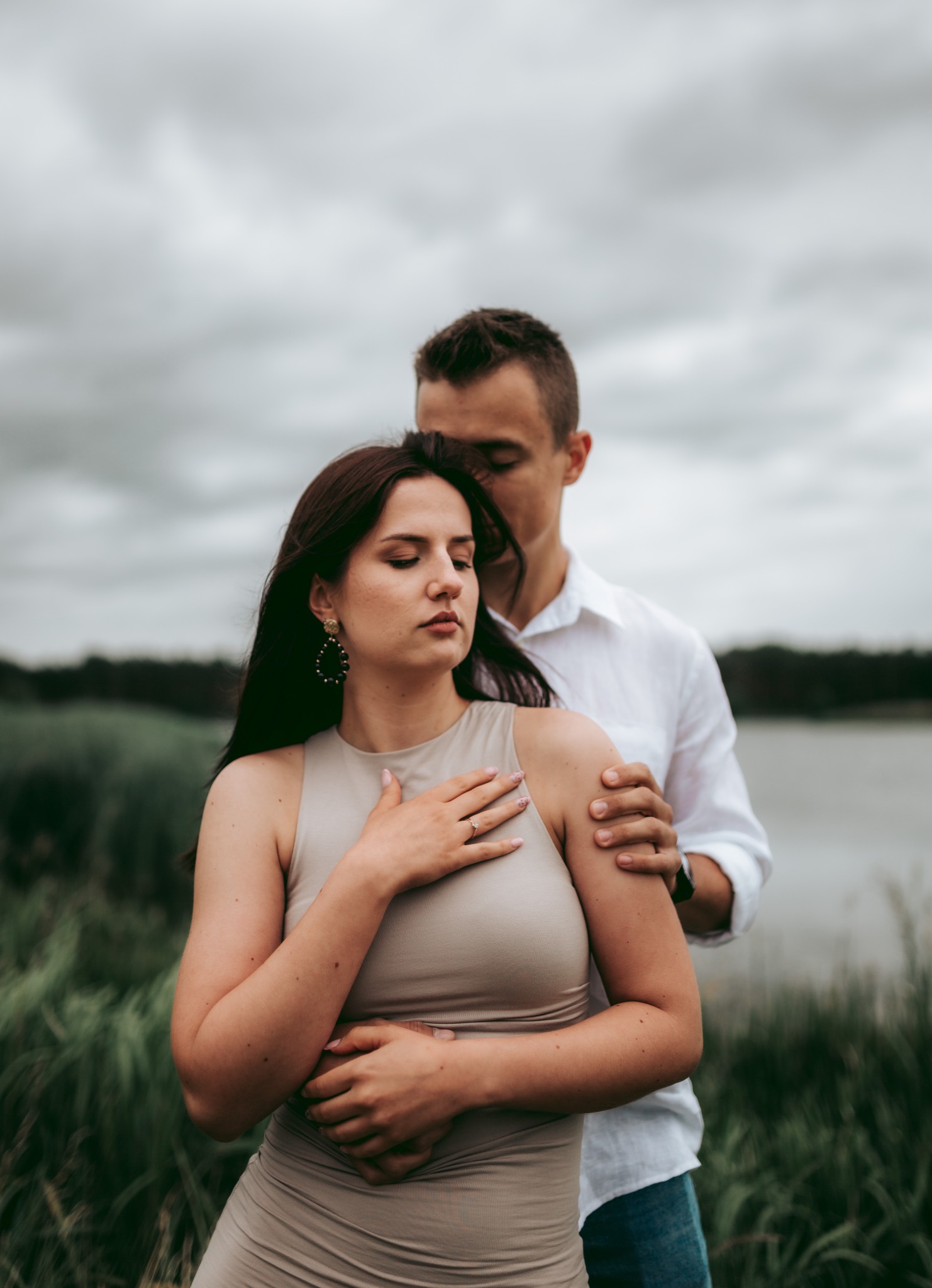 Młoda para w objęciach na tle pochmurnego nieba i jeziora; kobieta w beżowej sukience z dużymi kolczykami i pierścionkiem zaręczynowym, mężczyzna w białej koszuli obejmuje ją od tyłu.