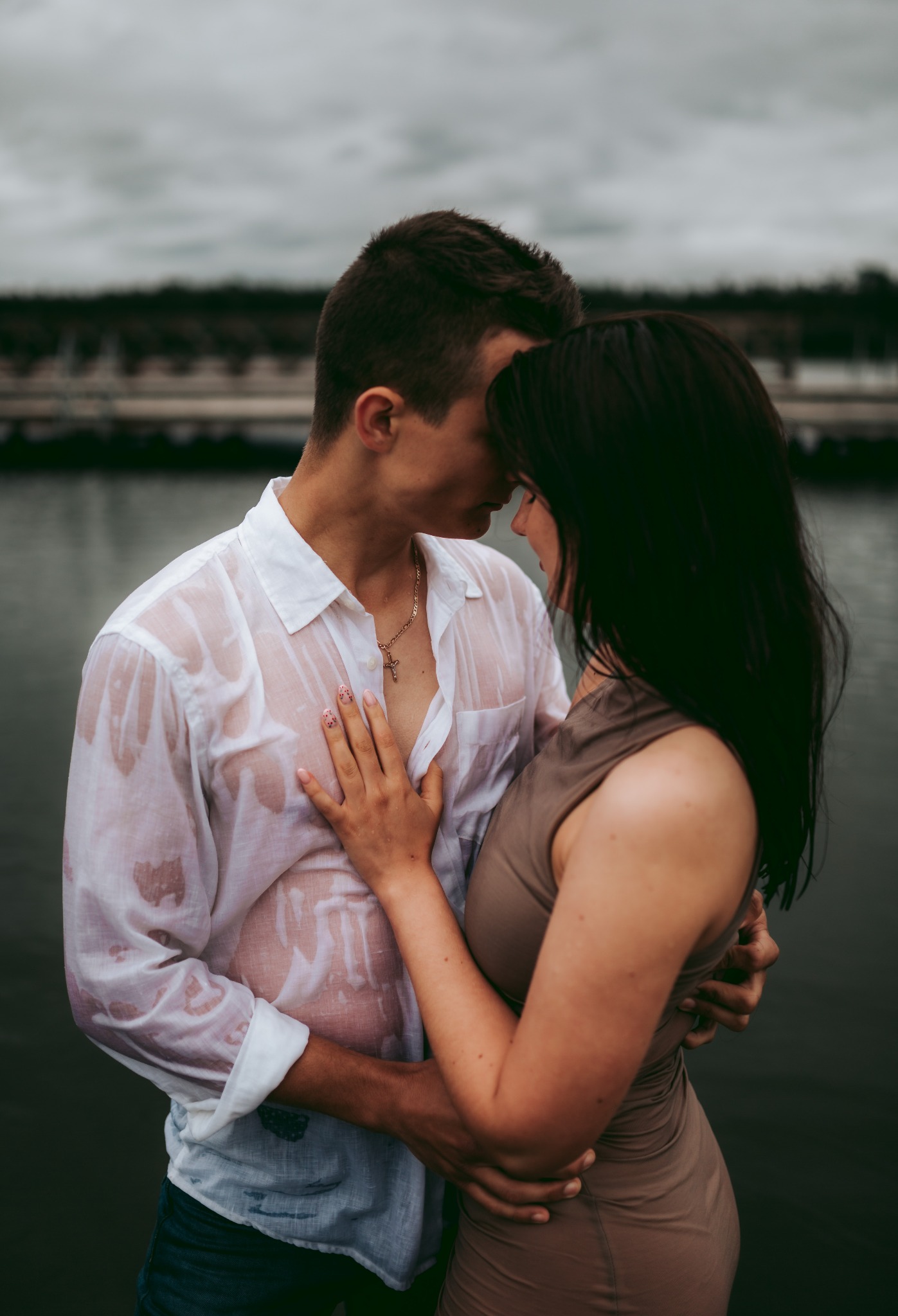Młoda para w mokrych ubraniach, obejmująca się na tle wody i pochmurnego nieba; ona w dopasowanej sukience w kolorze taupe, on w rozpiętej białej koszuli, widoczny złoty łańcuszek z krzyżykiem.