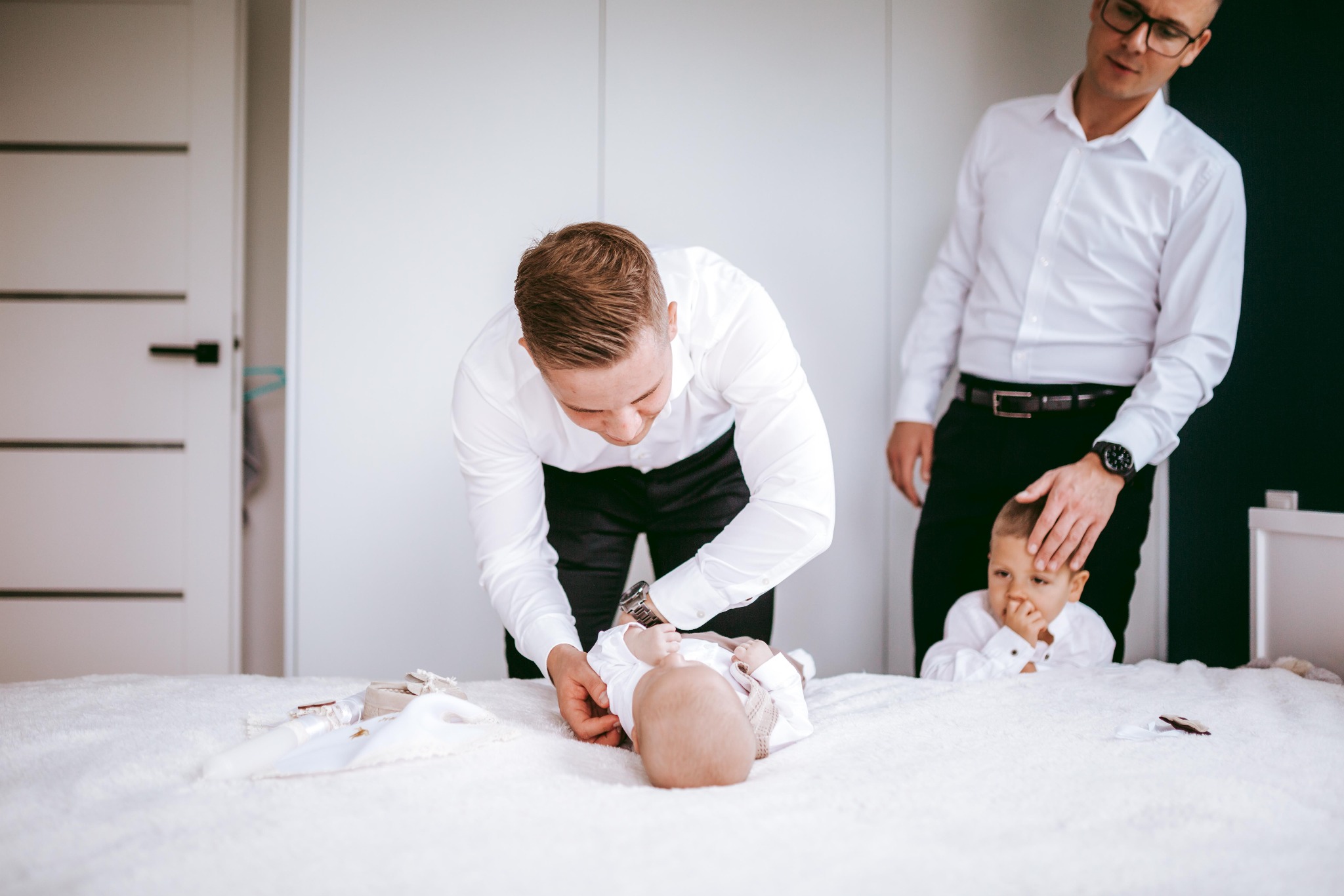 Dwóch mężczyzn w białych koszulach i czarnych spodniach przygotowuje niemowlę do chrztu na białym łóżku, obok leżą białe akcesoria do chrztu.