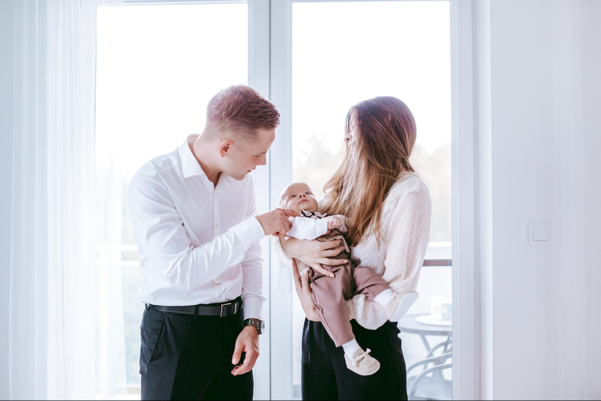 Elegancko ubrana rodzina: ojciec poprawia muchę niemowlęciu trzymanemu przez matkę na tle jasnego okna balkonowego.