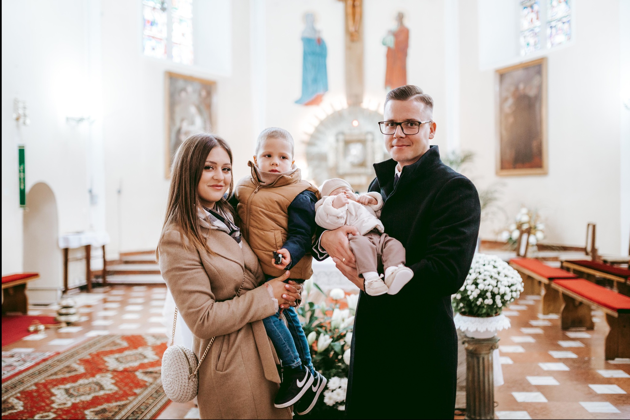 Rodzina z dwójką dzieci, w tym niemowlę w białym ubranku, pozuje w kościele na tle ołtarza i witraży.
