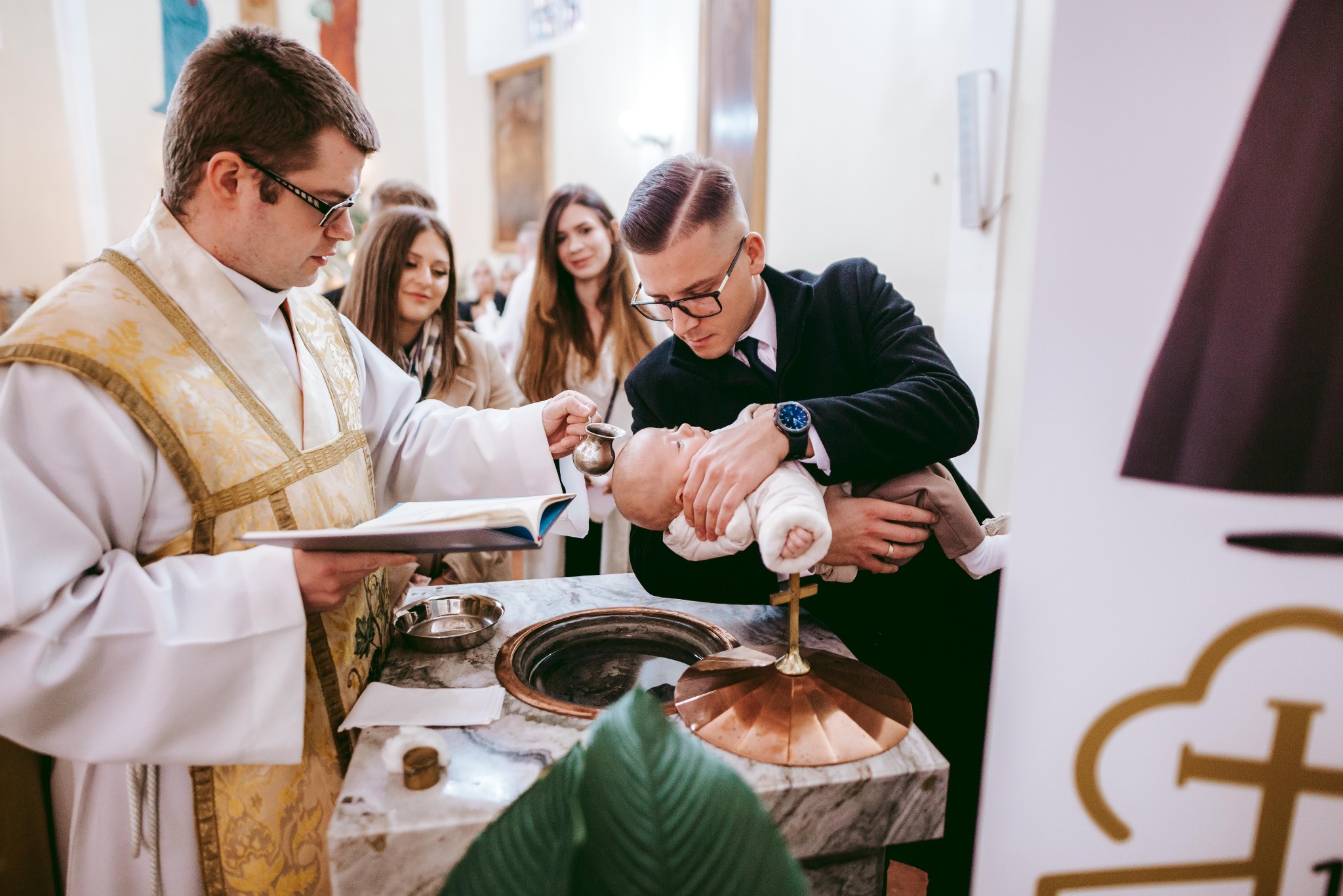 Ksiądz w ornacie polewa wodą główkę niemowlęcia trzymanego przez ojca w garniturze, w tle obserwują kobiety.