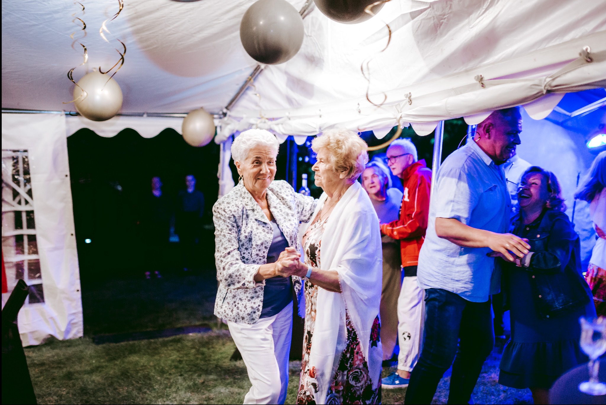 Dwie uśmiechnięte starsze kobiety tańczące pod białym namiotem z szarymi balonami, widoczni inni goście w tle.