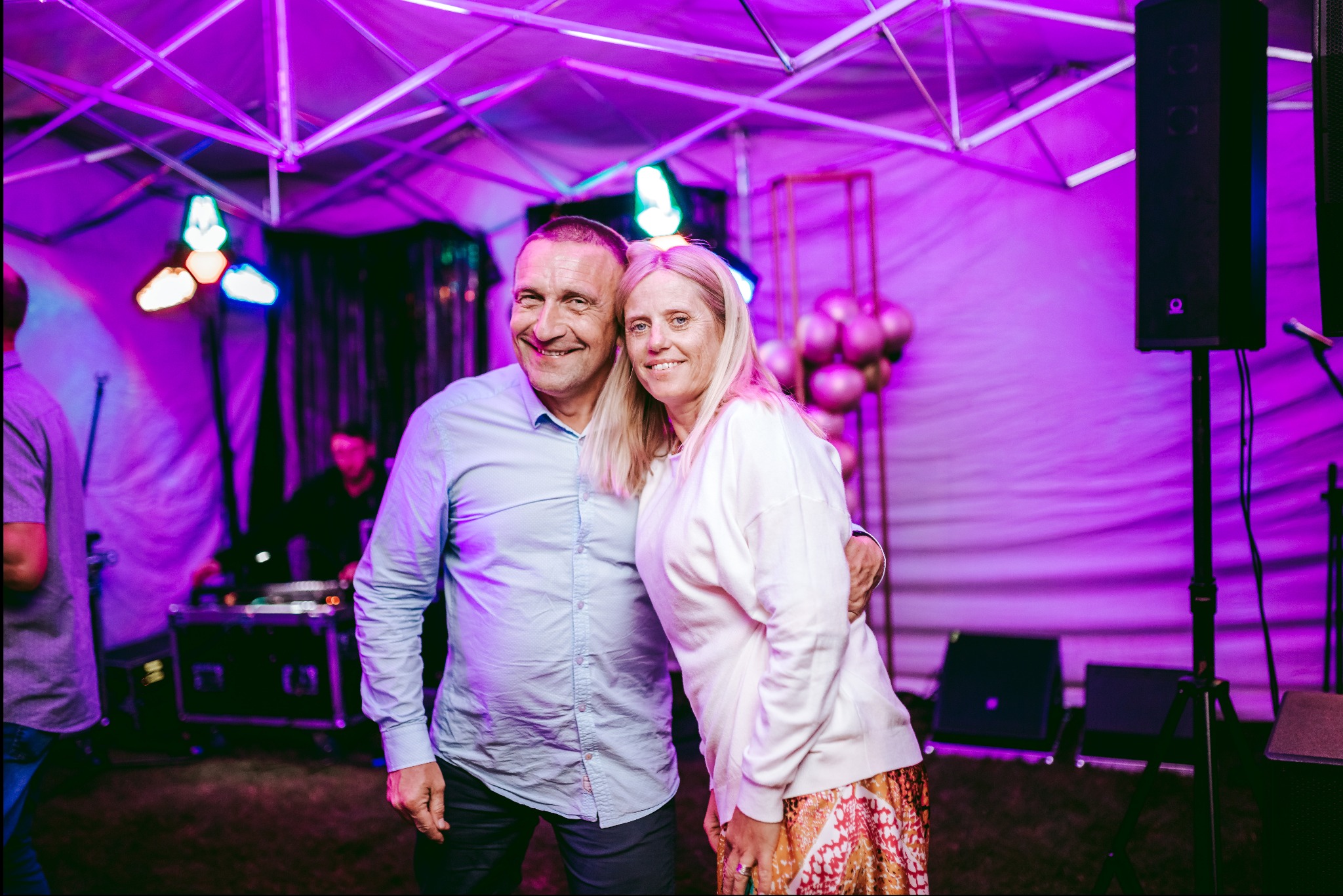 Para uśmiechnięta i obejmująca się na tle sceny z DJ-em, oświetleniem i dekoracjami w odcieniach fioletu.