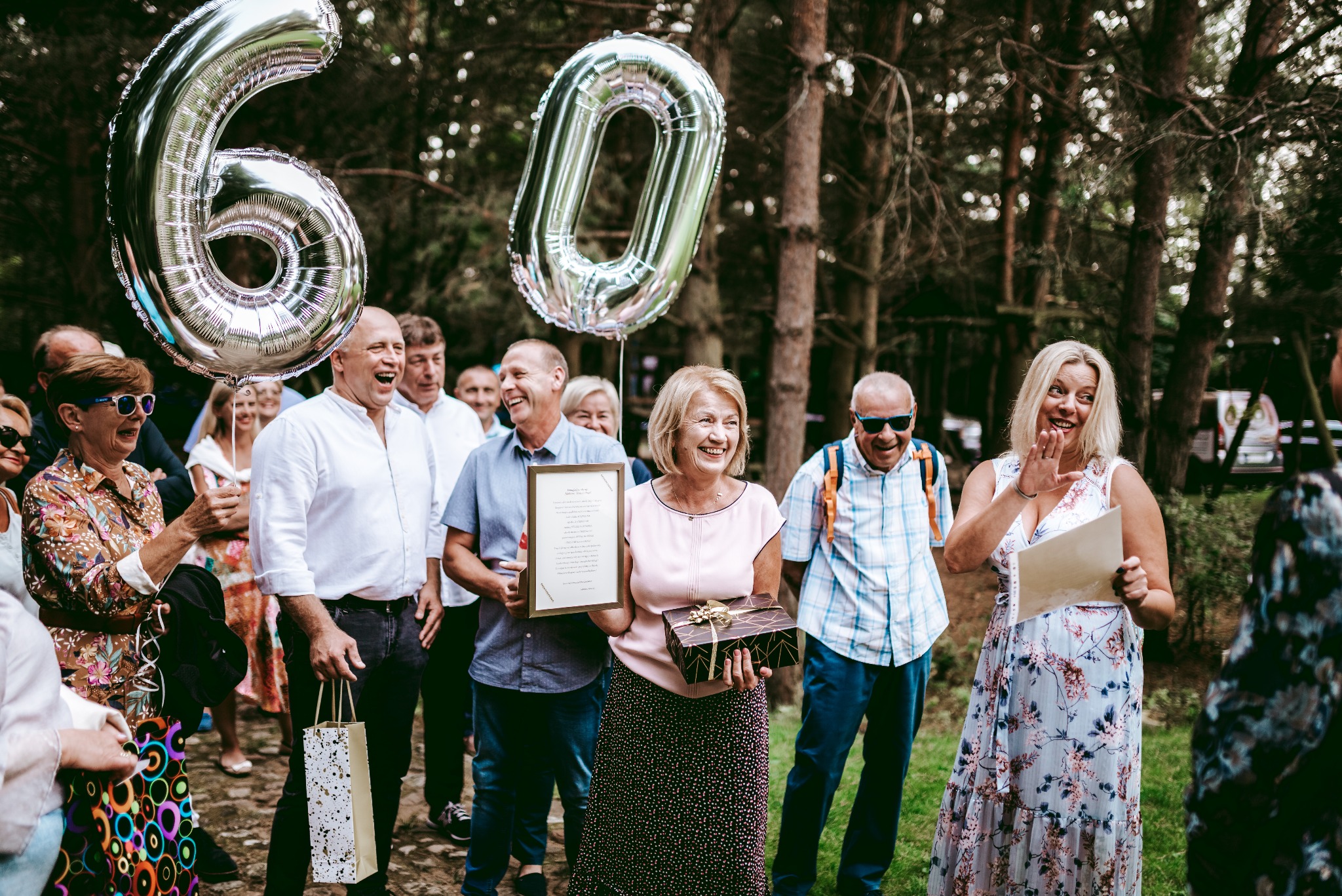 Uroczystość w plenerze z balonami w kształcie cyfr 60, kobieta trzyma prezent i ramkę z tekstem, a inna osoba gestykuluje, otoczeni przez uśmiechniętych gości na tle drzew.