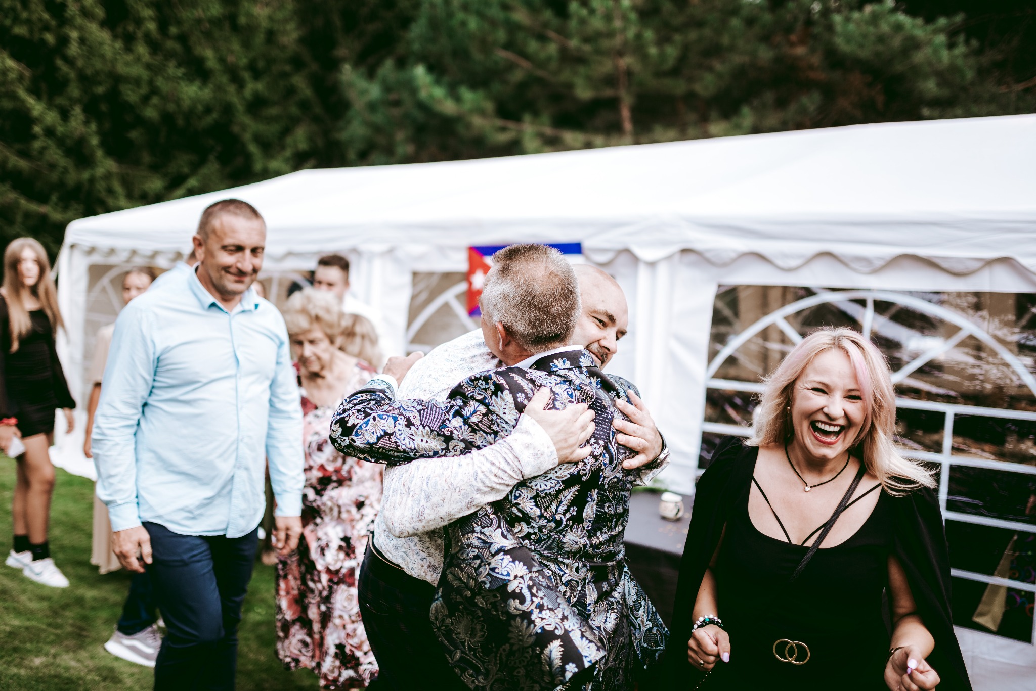 Uśmiechnięta kobieta z różowymi pasemkami na włosach, w czarnej sukience, śmieje się na tle białego namiotu i grupy ludzi, w tym dwóch mężczyzn obejmujących się w ozdobnym, wzorzystym garniturze.
