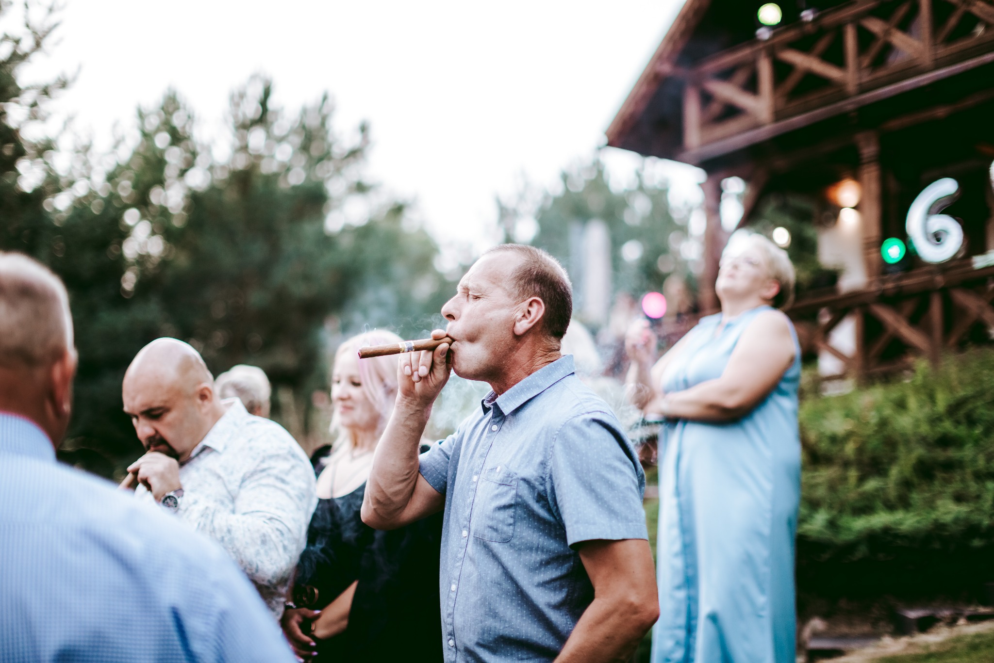 Mężczyzna w niebieskiej koszuli pali cygaro na tle drewnianej konstrukcji z dekoracyjnym oświetleniem i innymi osobami w tle.