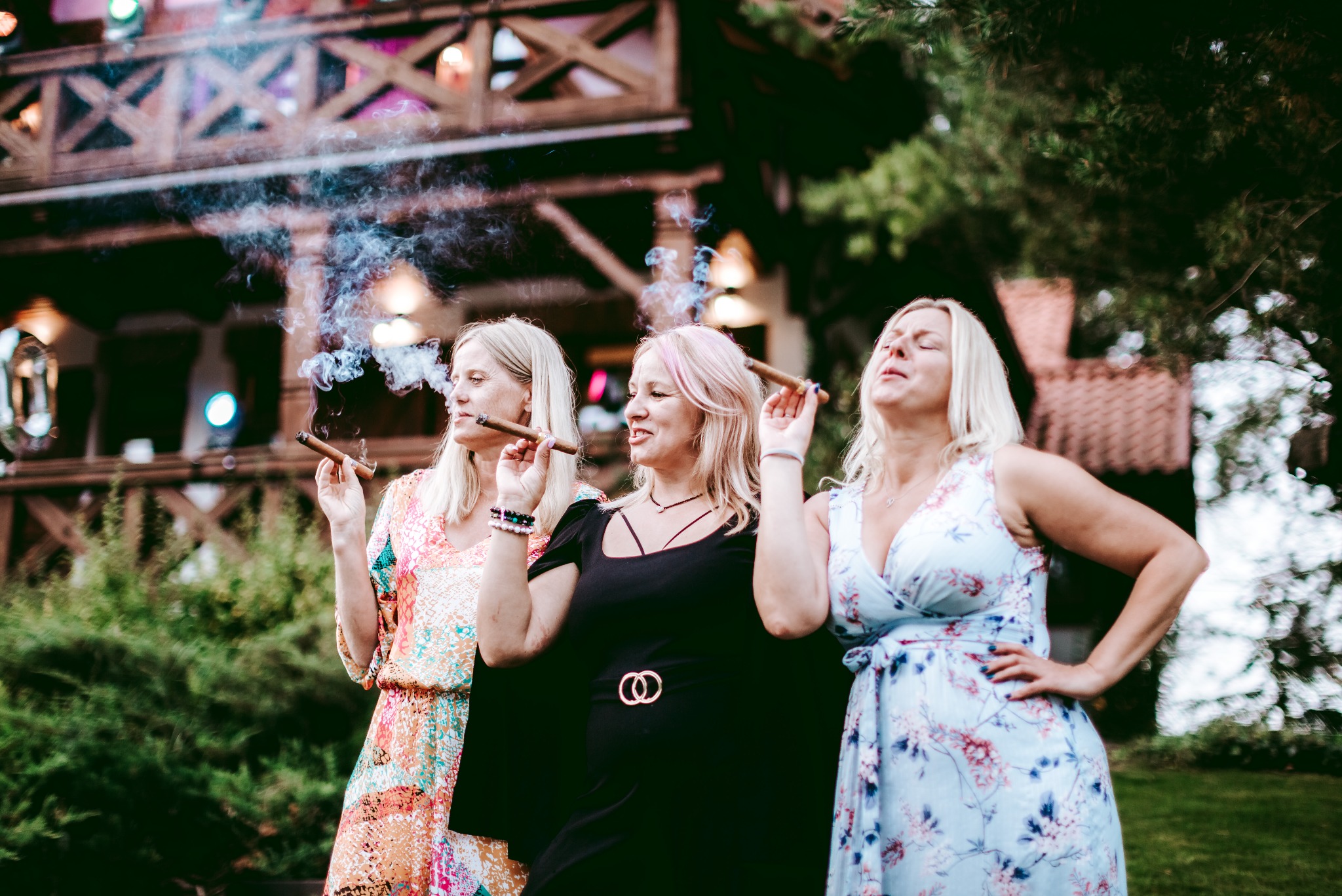 Trzy kobiety w sukienkach pozują z cygarami na tle drewnianej konstrukcji z oświetleniem i zieleni