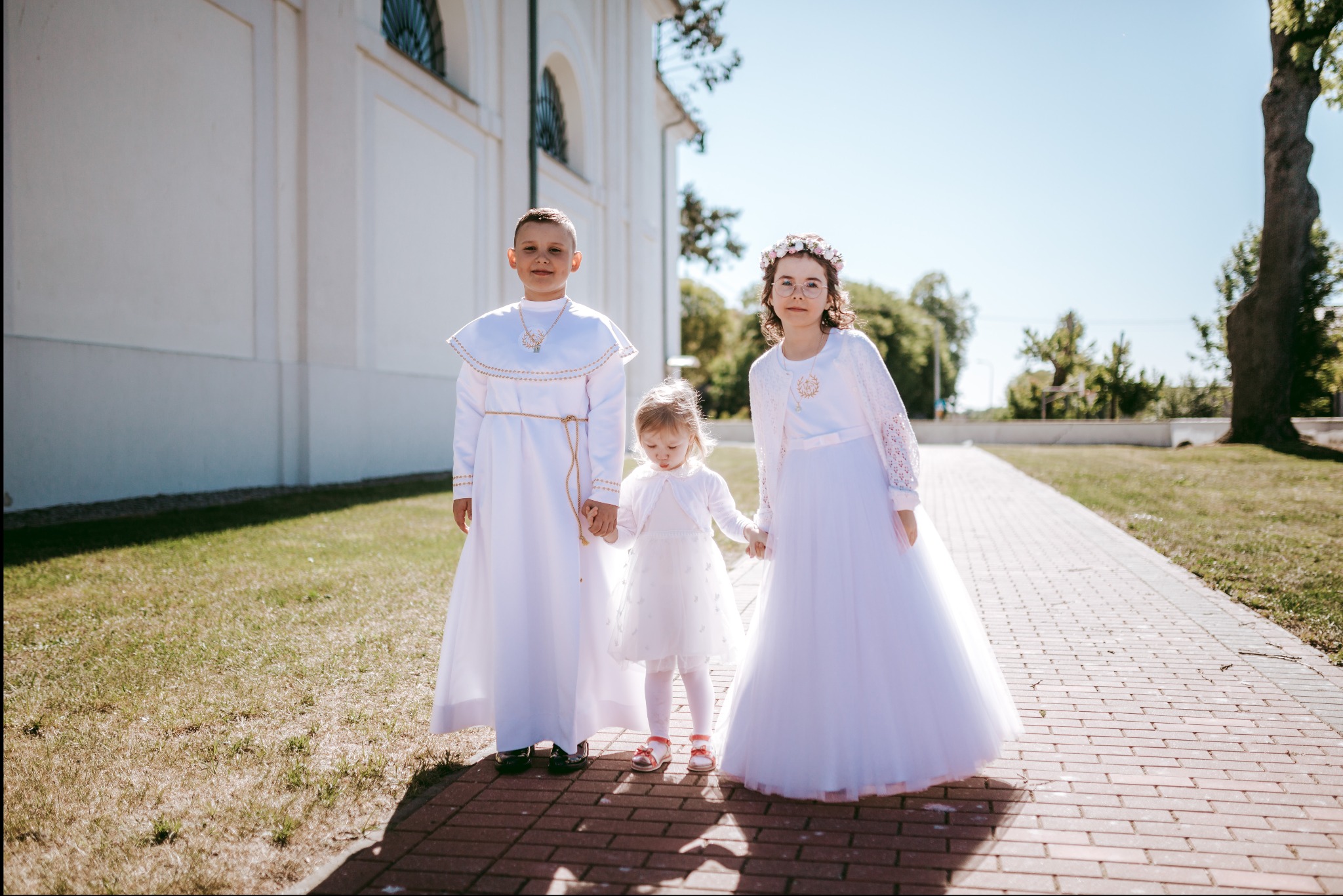 Troje dzieci w białych strojach komunijnych pozuje na tle kościoła w słoneczny dzień. Chłopiec z krótkimi włosami, dziewczynka w okularach z wiankiem na głowie i mała dziewczynka trzymają się za...
