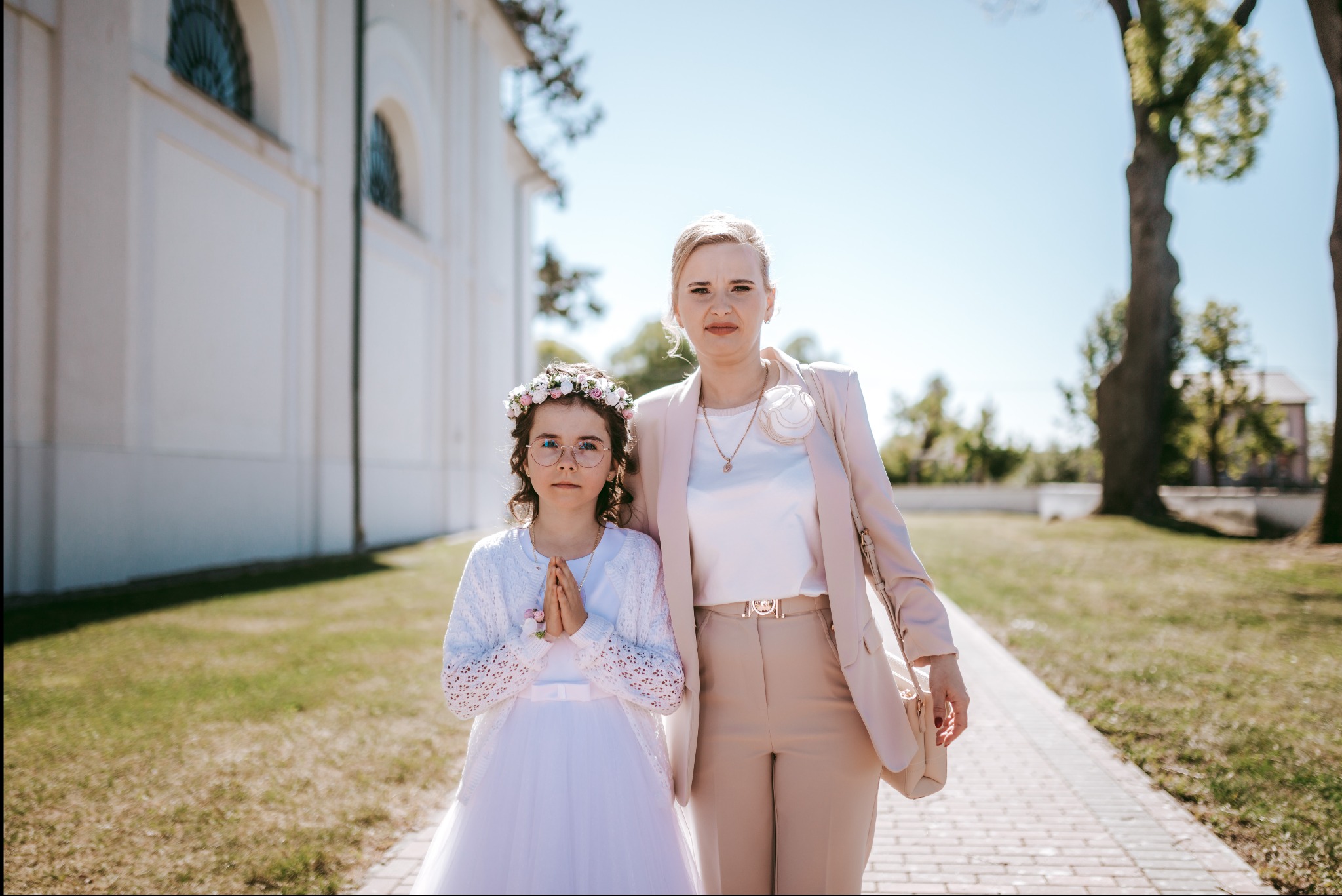 Dziewczynka w białej sukience komunijnej z wiankiem na głowie, stojąca obok kobiety w beżowym garniturze, na tle białego budynku i drzew
