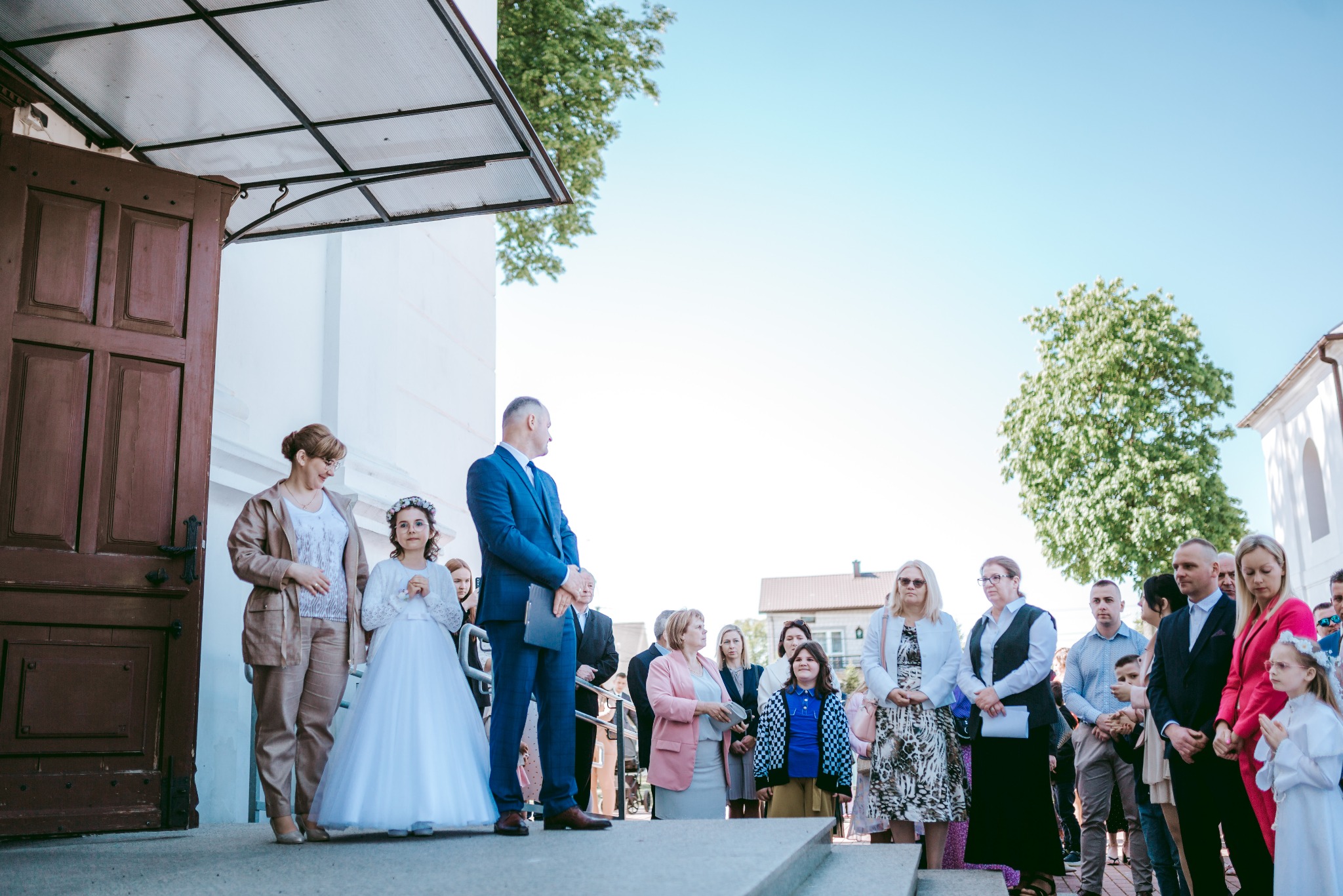 Grupa osób, w tym dziewczynka w białej sukience i wianku, stoi na schodach przed budynkiem z drewnianymi drzwiami podczas uroczystości.