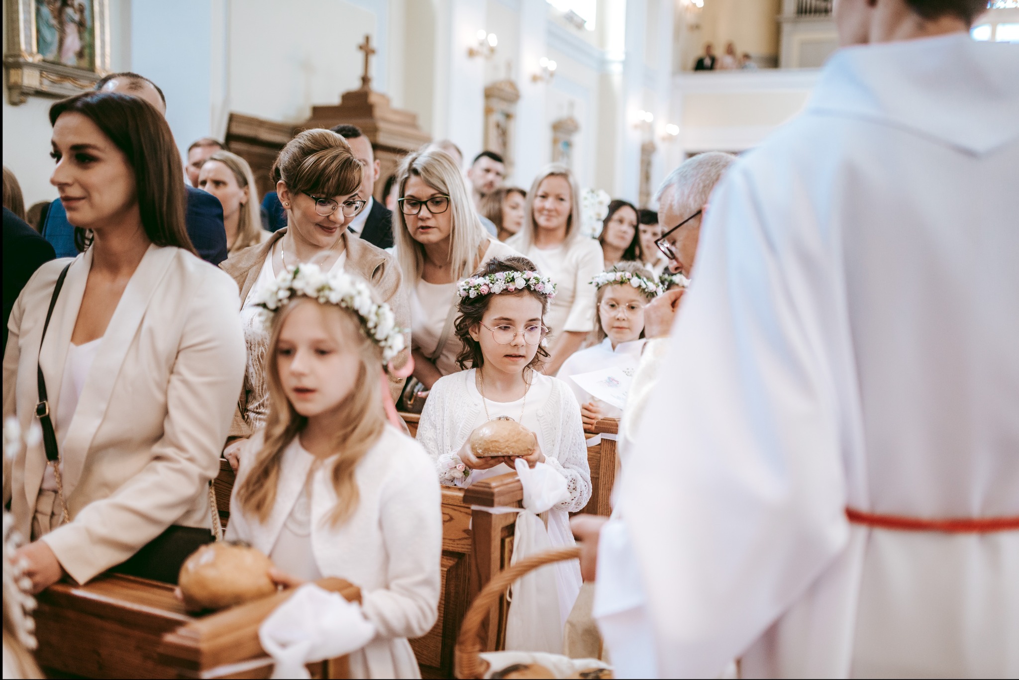 Uczestnicy Pierwszej Komunii Świętej w kościele, dziewczynki w wiankach trzymają chleb, obecny ksiądz w białej szacie z czerwonym pasem.