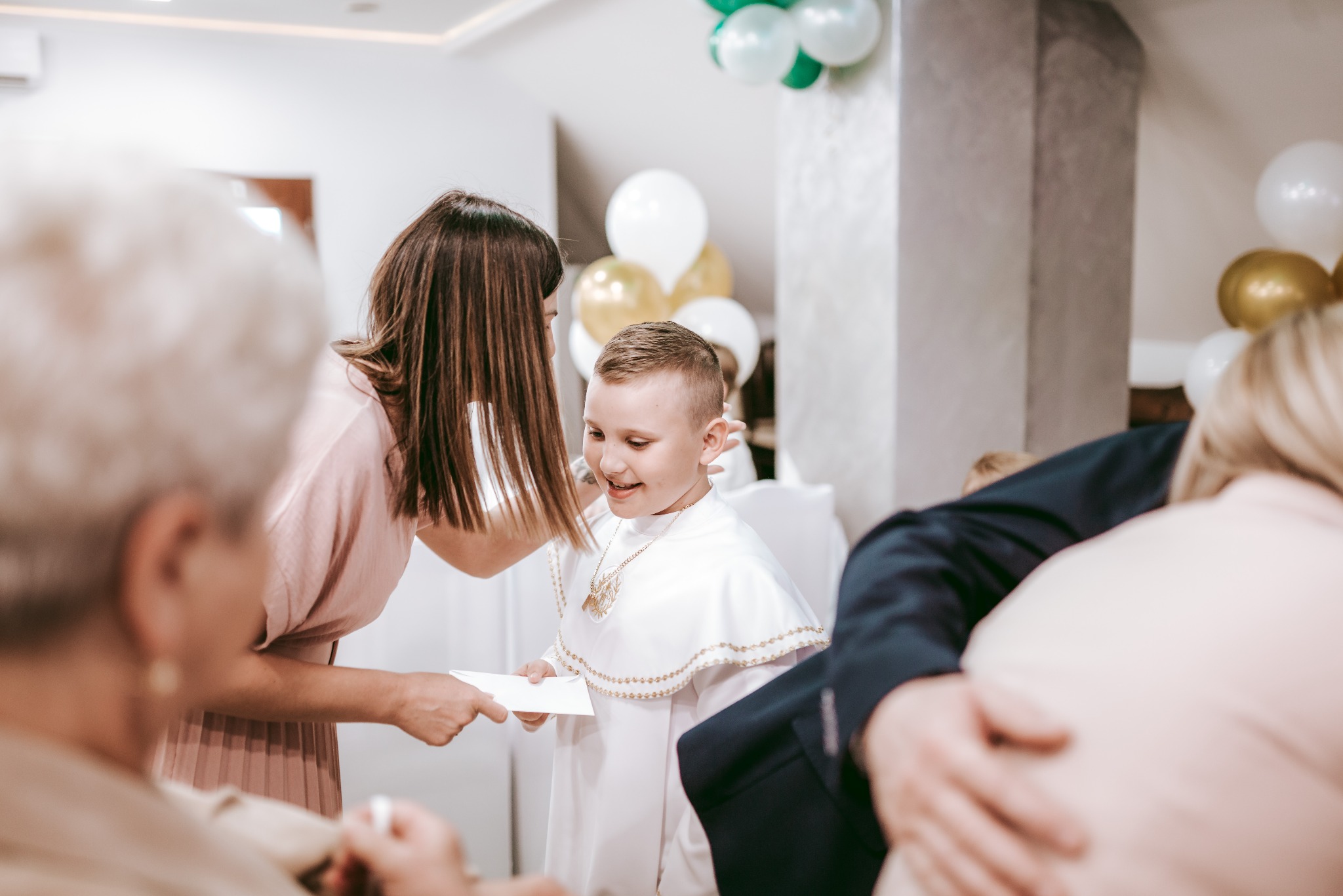 Chłopiec w białej albie z dekoracyjnym złotym haftem, uśmiechnięty, otrzymuje kartkę od kobiety w różowej sukience, w tle balony.