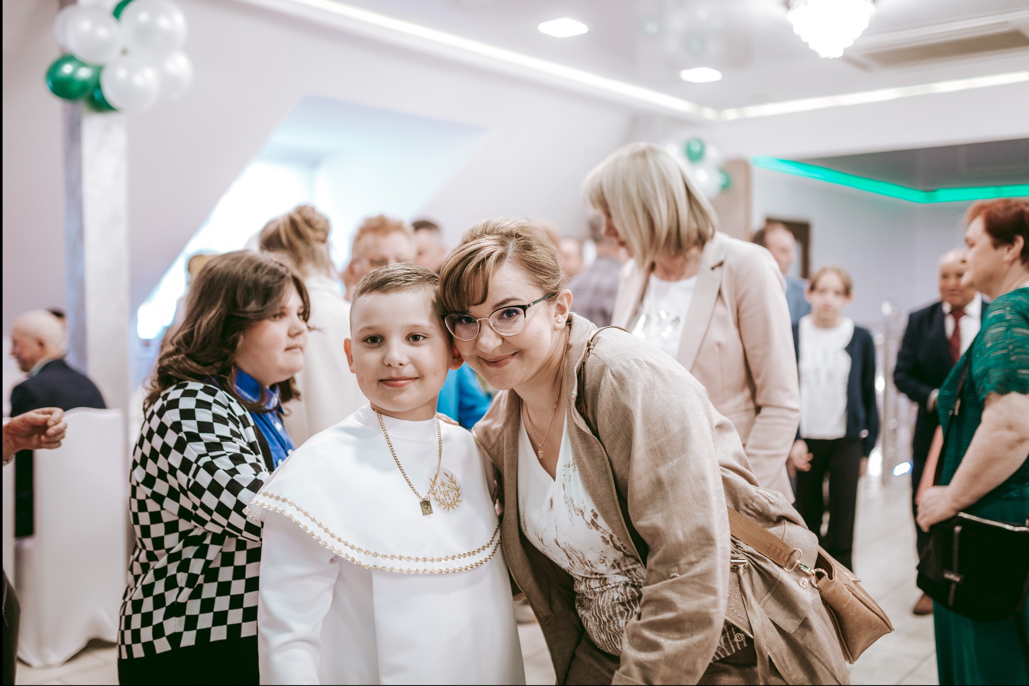 Chłopiec w stroju komunijnym pozuje z uśmiechniętą kobietą na tle gości i dekoracji z białych i zielonych balonów.