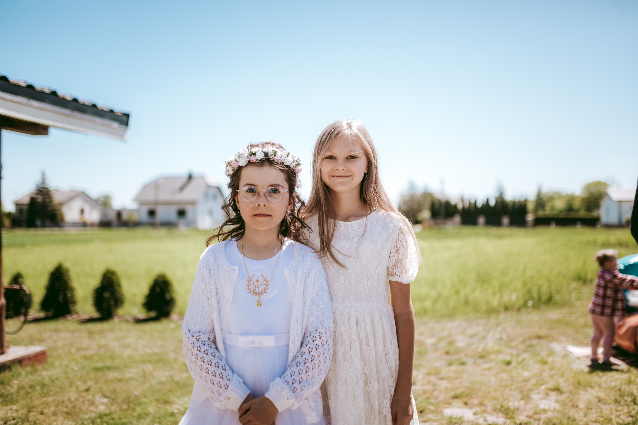 Dwie dziewczynki w białych sukienkach, jedna z wiankiem na głowie i okularami, druga z delikatnym uśmiechem, na tle zielonego trawnika i zabudowań w słoneczny dzień.