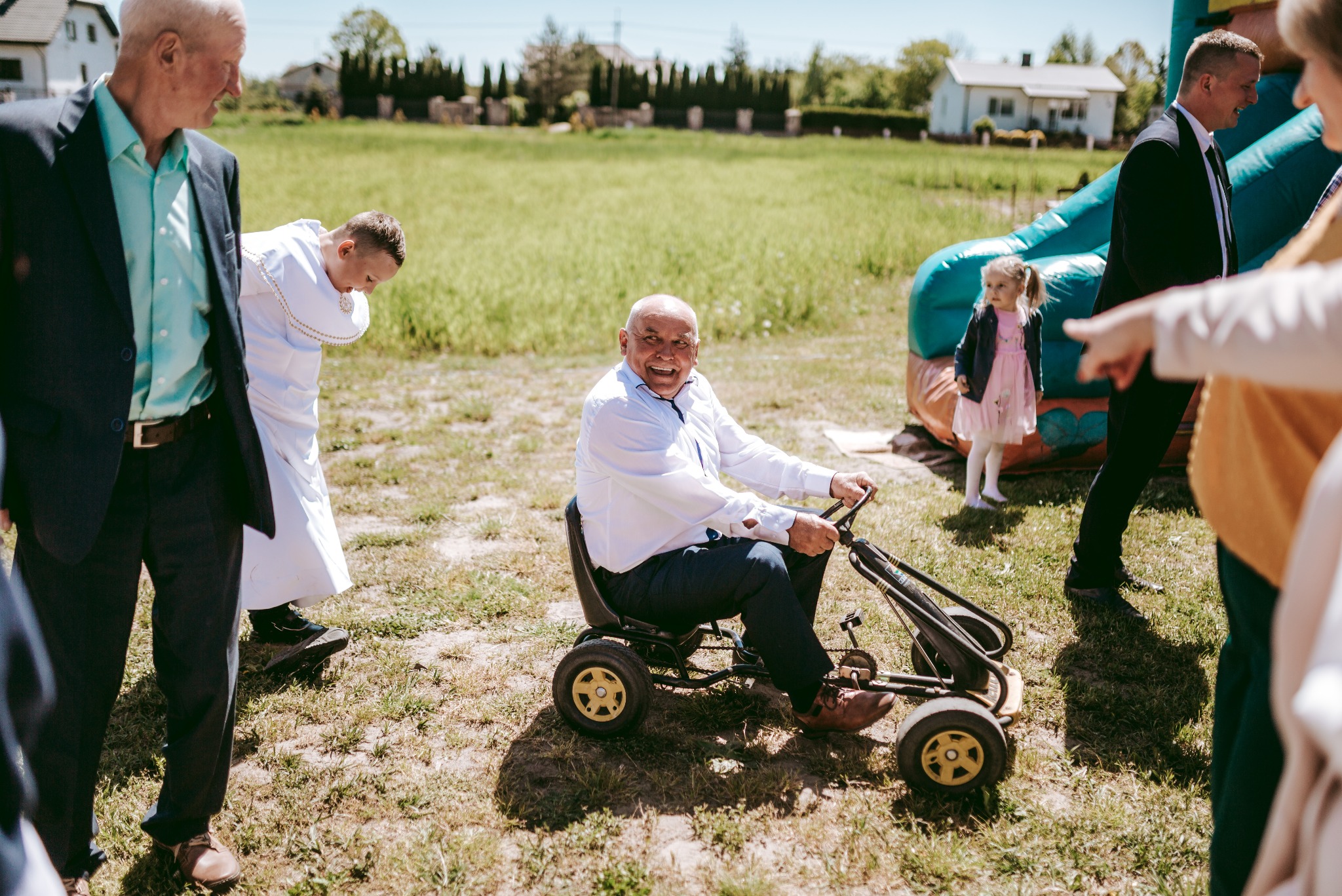 Uśmiechnięty mężczyzna w białej koszuli jedzie na gokarcie po trawiastym terenie podczas uroczystości, w tle dmuchana zjeżdżalnia, dziecko w różowej sukience i osoby w eleganckich strojach.