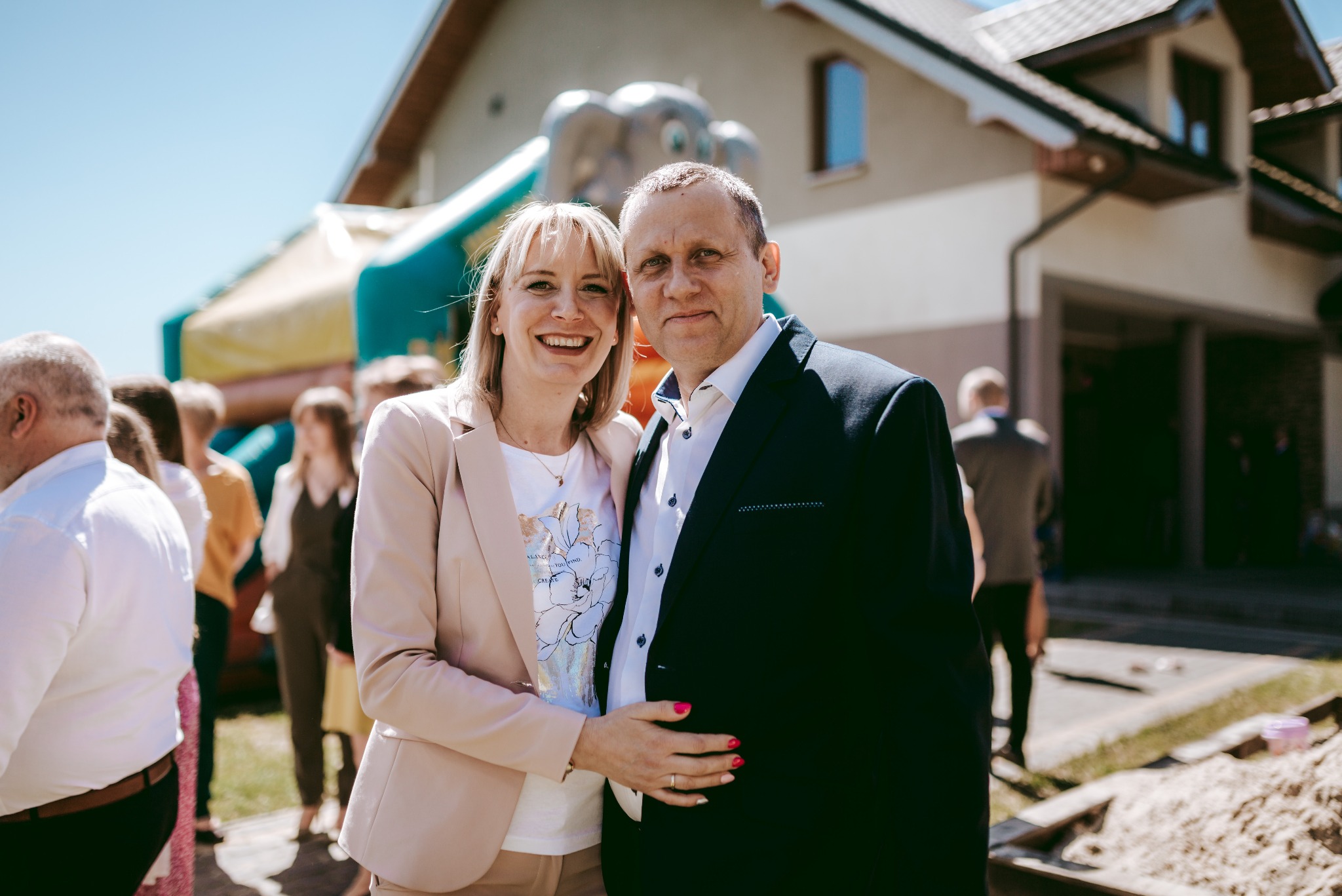 Uśmiechnięta para w eleganckich strojach pozuje na tle domu z dmuchanym zamkiem w tle, widoczni również goście.