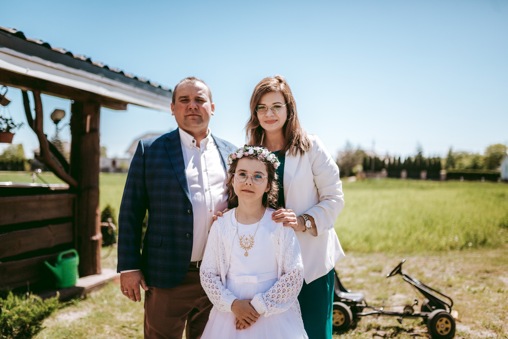 Rodzinne zdjęcie portretowe dziewczynki w białej sukience komunijnej i wianku, pozującej z rodzicami na tle ogrodu w słoneczny dzień. W tle widoczny wózek na pedały.