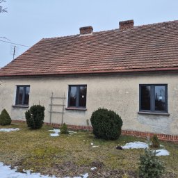 BramAD Adrian Kulik - Dom jednorodzinny z trzema ciemnymi oknami, drabina oparta o ścianę, ogródek z iglakami i resztkami śniegu, dach z czerwonej dachówki.