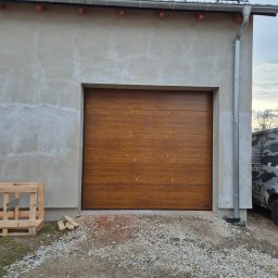 BramAD Adrian Kulik - Nowa brama garażowa segmentowa w kolorze drewna zamontowana w surowym budynku, widoczny fragment kamuflażowego samochodu dostawczego i paleta obok.