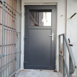 Szare drzwi zewnętrzne z niewielkim przeszkleniem w górnej części, zamontowane w wejściu do budynku, widoczne metalowe schody i krata zabezpieczająca.