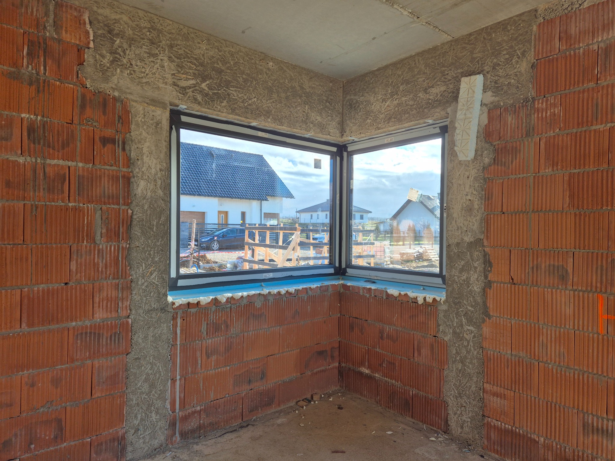 Narożne okno z czarną ramą, zamontowane w surowym murze z czerwonej cegły, widok na domy jednorodzinne i błękitne niebo