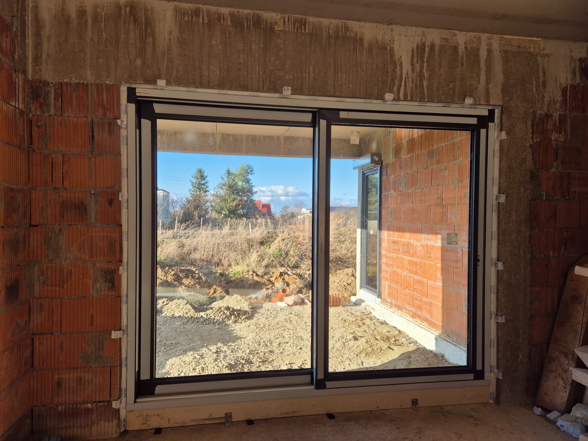 Dwuskrzydłowe okno przesuwne z widokiem na ogród w trakcie budowy, zamontowane w surowym murze z cegły.