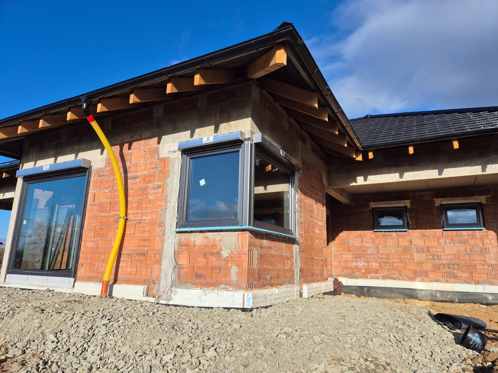 Surowy budynek z cegły z zamontowanymi oknami z szarymi ramami i roletami zewnętrznymi w kolorze aluminium, widoczna żółta rura wentylacyjna przy ścianie, dach pokryty czarną dachówką, teren wokół...