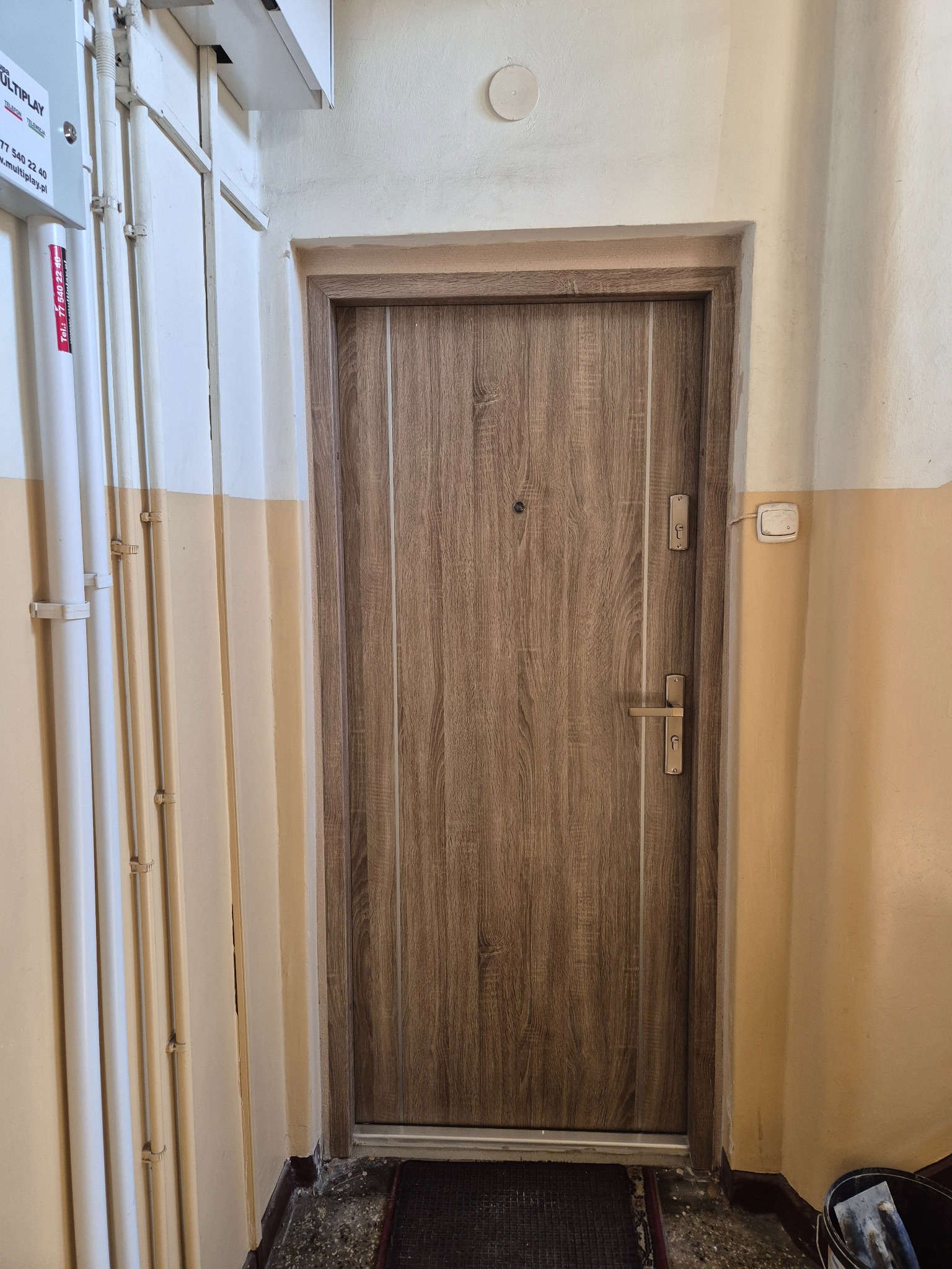 Brązowe drzwi z wizjerem i metalową klamką, zamontowane w klatce schodowej z żółto-białymi ścianami i widocznymi rurami instalacyjnymi.