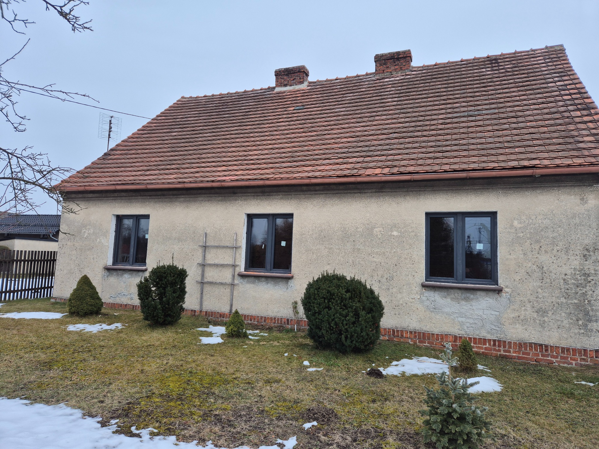 Dom jednorodzinny z trzema ciemnymi oknami, drabina oparta o ścianę, ogródek z iglakami i resztkami śniegu, dach z czerwonej dachówki.