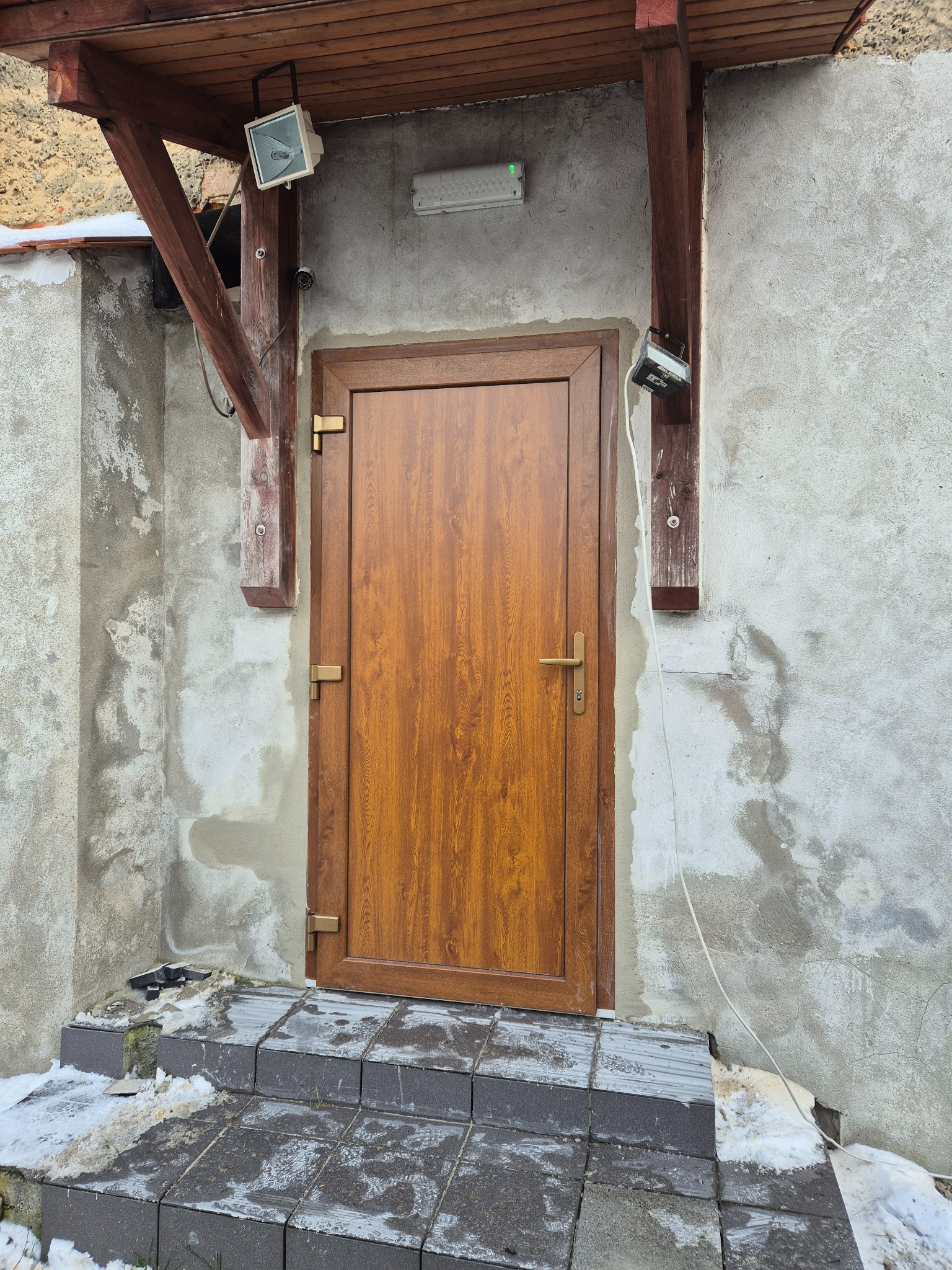 Brązowe drzwi zewnętrzne z ościeżnicą PCV, widoczne z zewnątrz domu z szarą elewacją, oświetlone dwoma reflektorami, z fragmentami śniegu na schodach.