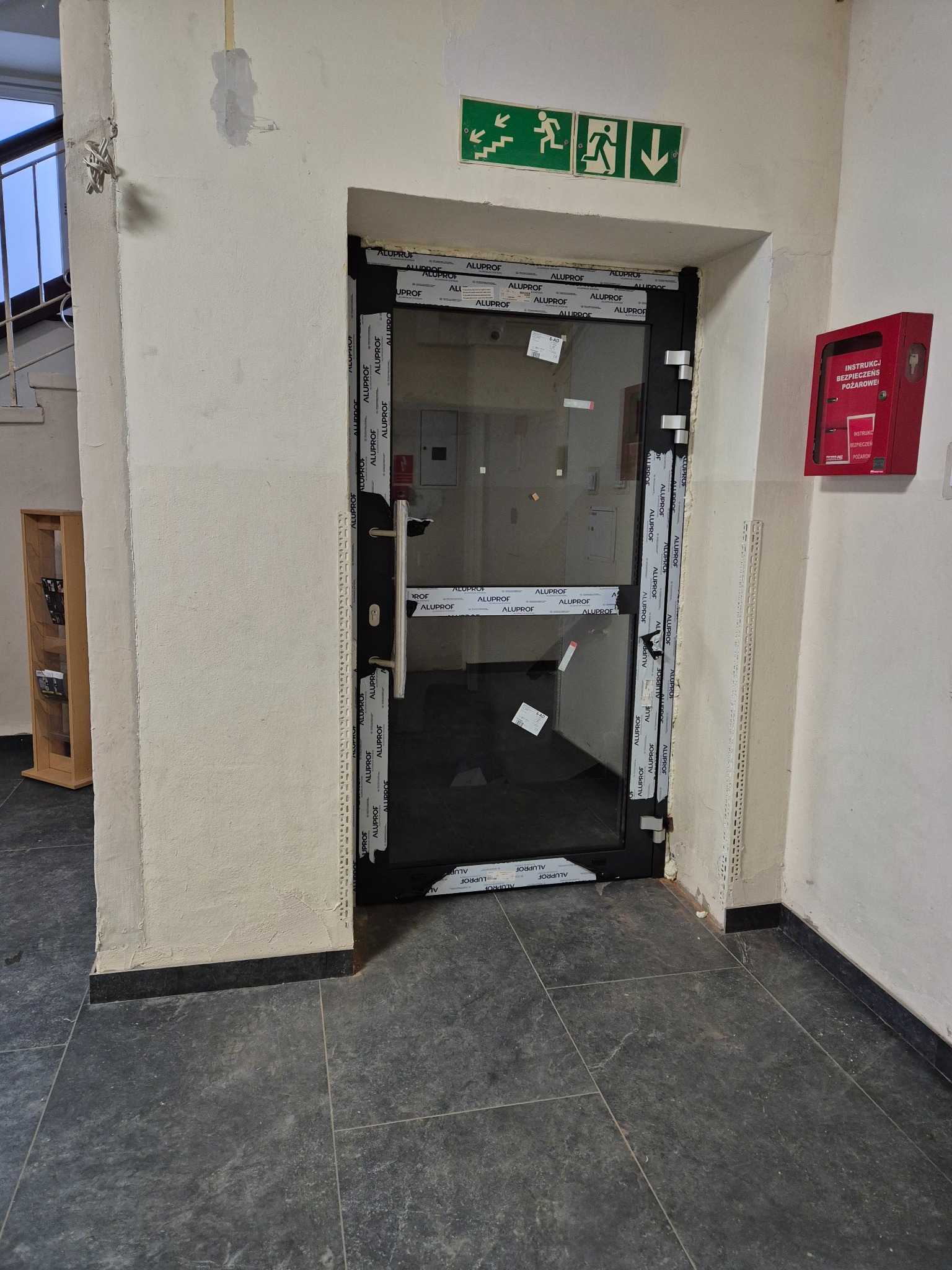 Ciemna, przeszklona konstrukcja drzwiowa z widocznymi oznaczeniami producenta na ramie, zainstalowana w korytarzu budynku z oznakowaniem ewakuacyjnym i skrzynką przeciwpożarową.