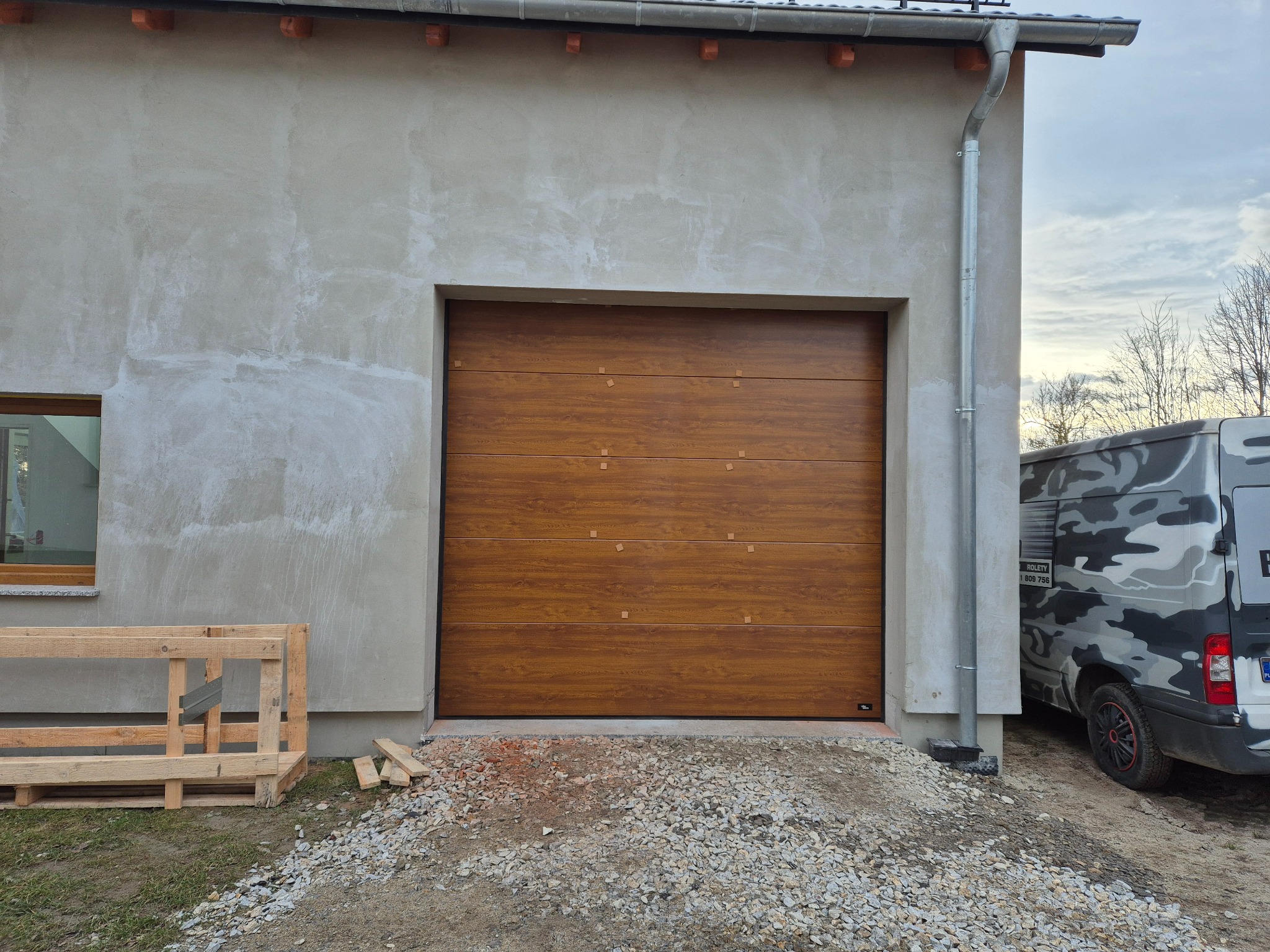 Nowa brama garażowa segmentowa w kolorze drewna zamontowana w surowym budynku, widoczny fragment kamuflażowego samochodu dostawczego i paleta obok.