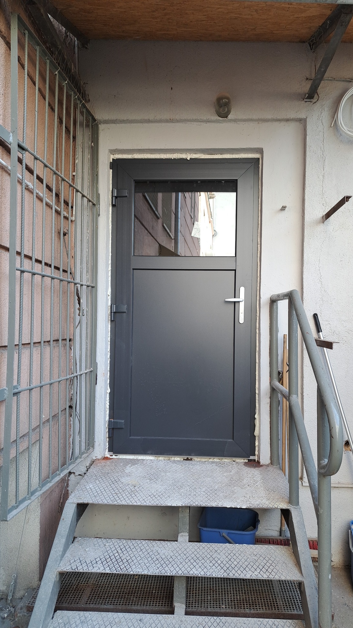 Szare drzwi zewnętrzne z niewielkim przeszkleniem w górnej części, zamontowane w wejściu do budynku, widoczne metalowe schody i krata zabezpieczająca.