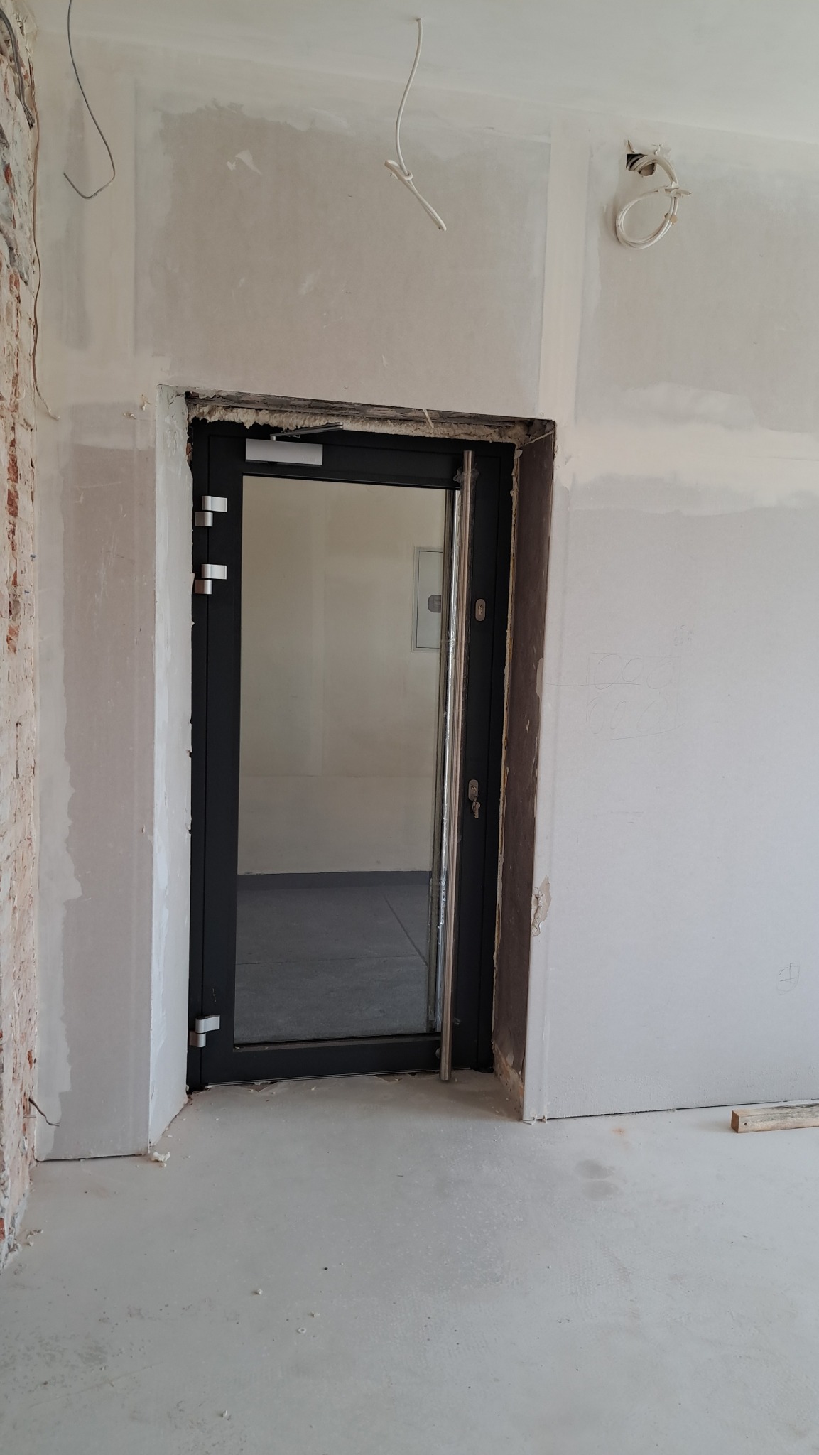 Ciemna, częściowo otwarta, przeszklona konstrukcja drzwiowa z widocznymi zamkami, osadzona w surowym otworze ściany w trakcie remontu, z odsłoniętą cegłą po lewej stronie i widocznymi przewodami...