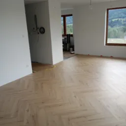 Salon z nowo ułożoną podłogą z paneli w jodełkę, widok na góry za oknem, niedokończone instalacje elektryczne na ścianie.