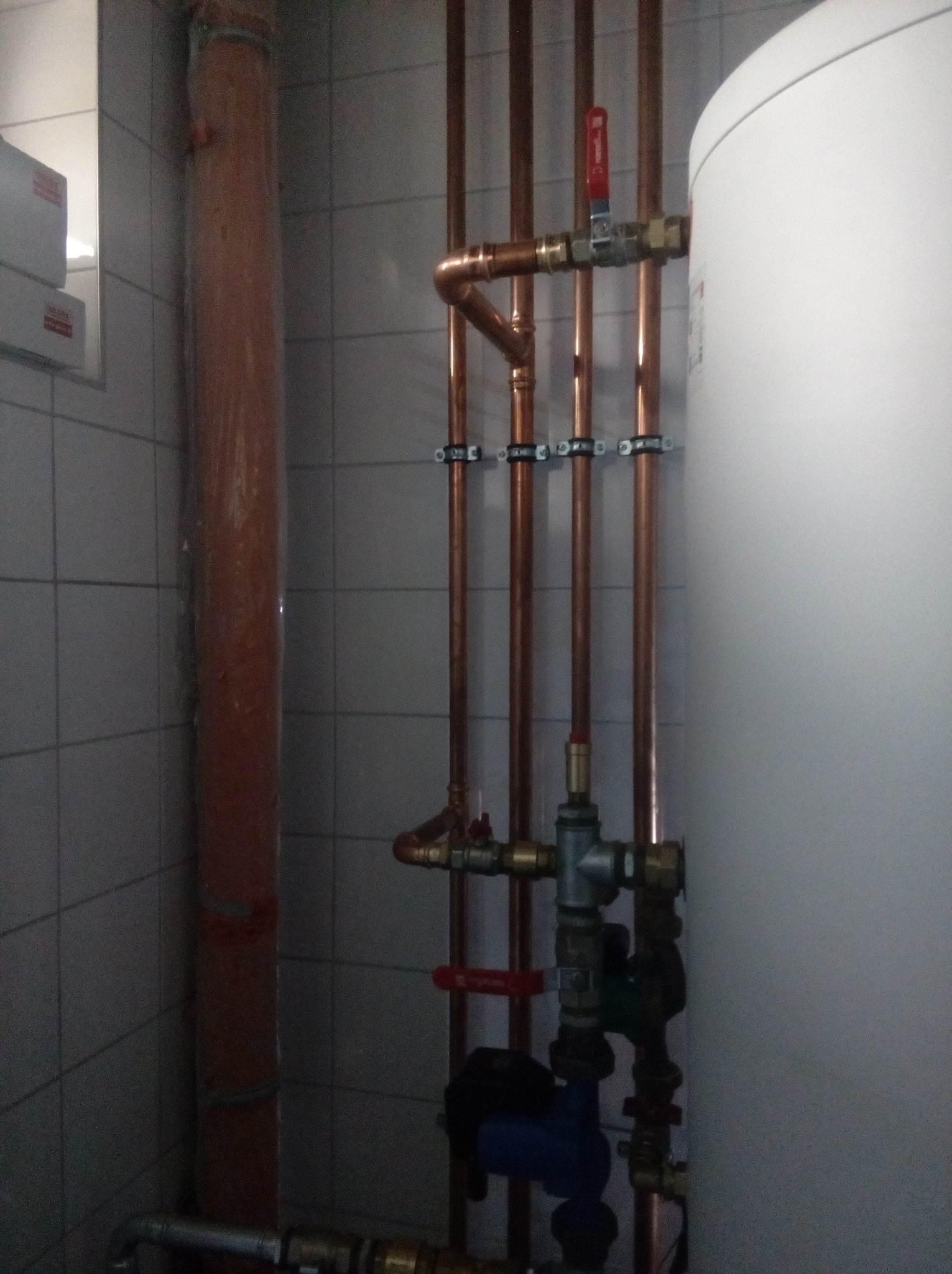 Miedziane rury instalacji wodnej z zaworami i pompą obiegową, przymocowane do ściany z szarych płytek, obok biały zasobnik ciepłej wody.