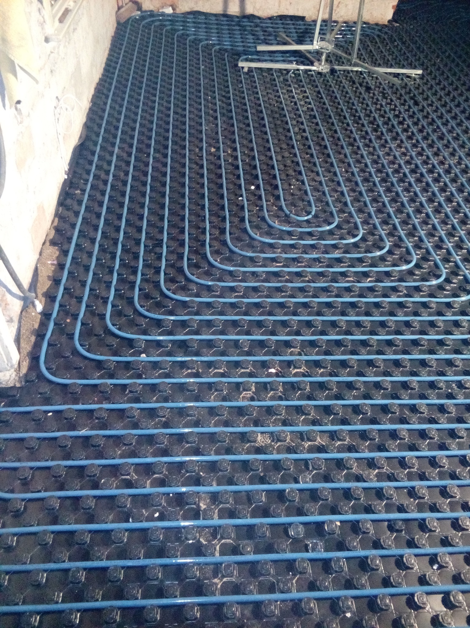 Niebieskie rury ogrzewania podłogowego ułożone na czarnej macie izolacyjnej z wypustkami, widoczne elementy montażowe w tle, surowy stan wykończenia pomieszczenia.