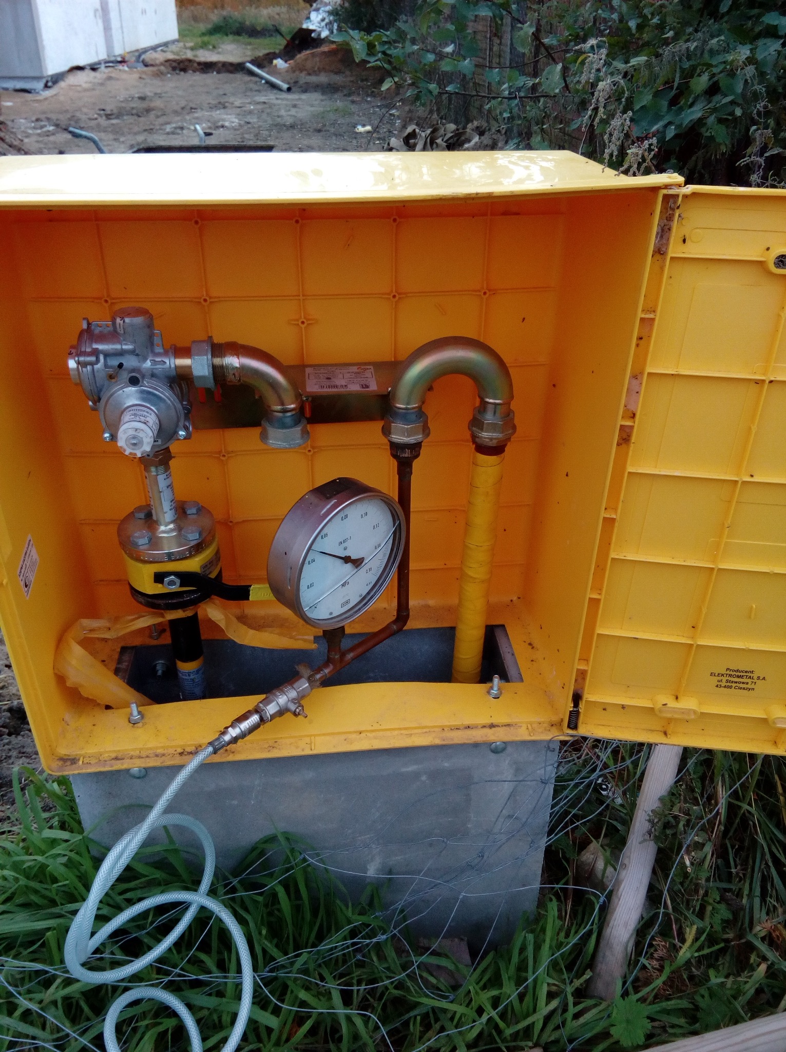 Żółta skrzynka gazowa z zainstalowanym reduktorem, manometrem i przewodami, widoczne elementy instalacji gazowej w otoczeniu trawy i ziemi.