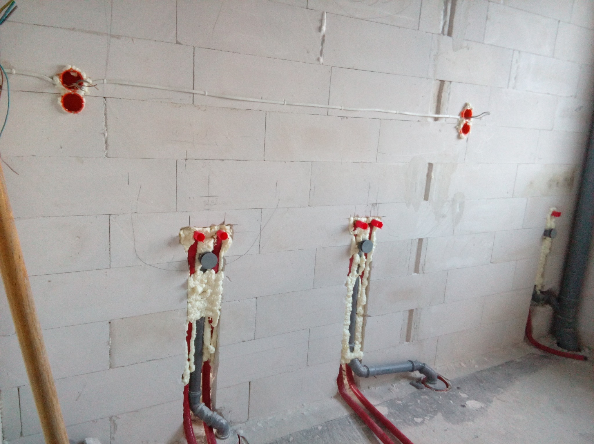 Montaż instalacji wodnej w bloku z betonu komórkowego, rury w kolorze czerwonym i szarym, zabezpieczone pianką montażową, widoczne puszki elektryczne zalane czerwoną farbą.
